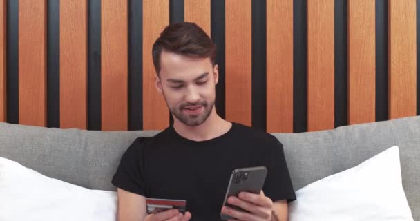 Jongeman zit op het bed en betaalt online aankopen met een smartphone, voert creditcardgegevens in op het scherm smartphone, online winkelen. — Stockvideo