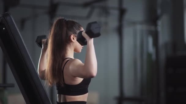 Entrenamiento femenino joven en el gimnasio, niña levanta pesas de mancuerna mientras está sentado en el banco, entrenamiento muscular con pesas. — Vídeos de Stock