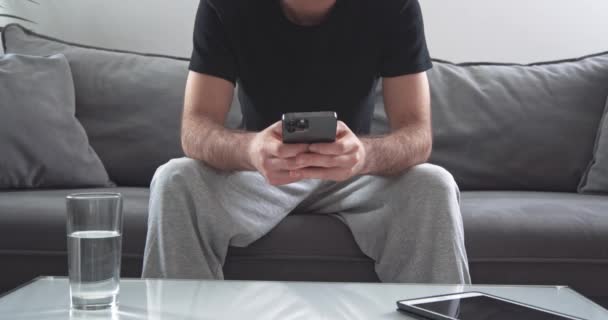Homem anônimo sentado no sofá na sala de estar e usa um smartphone moderno, um homem está digitando em um telefone celular, conversando online. — Vídeo de Stock