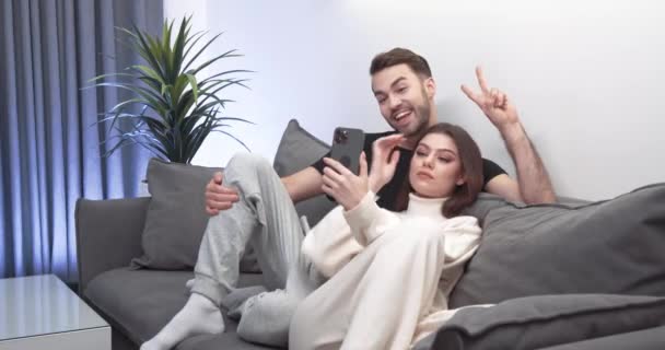Νεαρό ζευγάρι παίρνει μια selfie σε ένα smartphone, ένας άντρας και ένα κορίτσι κάθονται στον καναπέ στο σαλόνι και πυροβολούν ένα βίντεο σε ένα κινητό τηλέφωνο, αυτο-απομόνωση. — Αρχείο Βίντεο