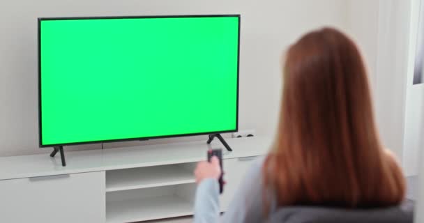 Menina está sentada no sofá na sala de estar e assistindo TV, tela verde, a menina muda de canais usando o controle remoto, modelo de chave chroma. — Vídeo de Stock