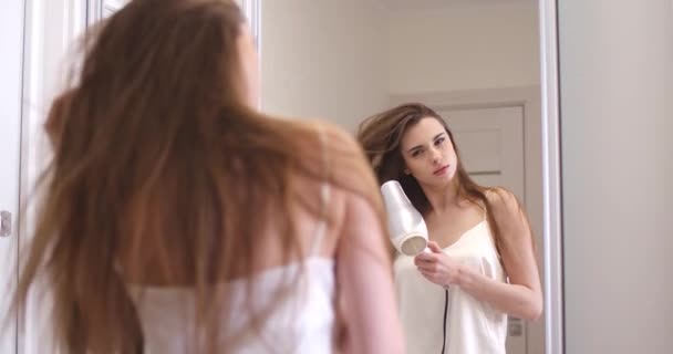Νεαρό όμορφο θηλυκό στέκεται κοντά στον καθρέφτη και στεγνώνει τα μαλλιά της με στεγνωτήρα μαλλιών, θέα της αντανάκλασης στον καθρέφτη, αργή κίνηση. — Αρχείο Βίντεο