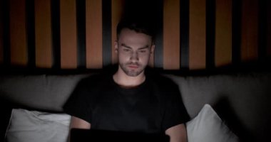 Genç adam yatakta uzanıyor ve dizüstü bilgisayarla çalışıyor, klavyeye mesaj yazıyor, geceleri çalışıyor, gece evden uzakta çalışıyor..