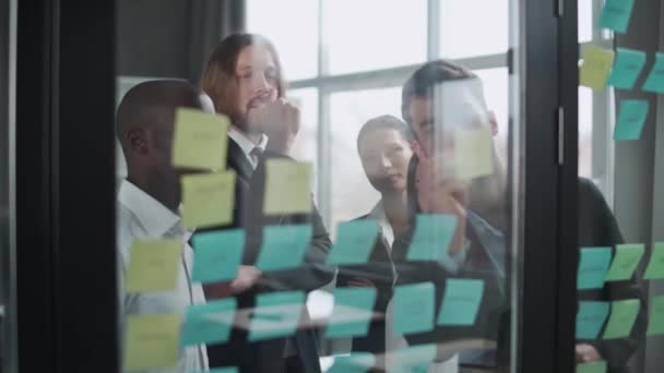 Business meeting, internationaal managementteam aan het werk, team staat naast een glazen bord met papieren stickers en notities, discussieert over werkplannen en financiële statistieken, brainstorm. — Stockvideo