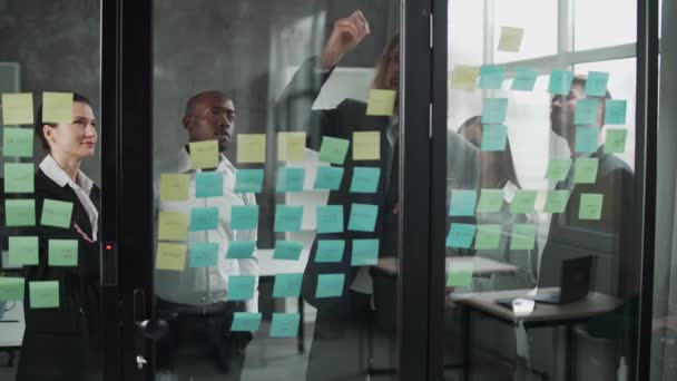 Business meeting, internationaal managementteam aan het werk, team staat naast een glazen bord met papieren stickers en notities, discussieert over werkplannen en financiële statistieken, brainstorm. — Stockvideo