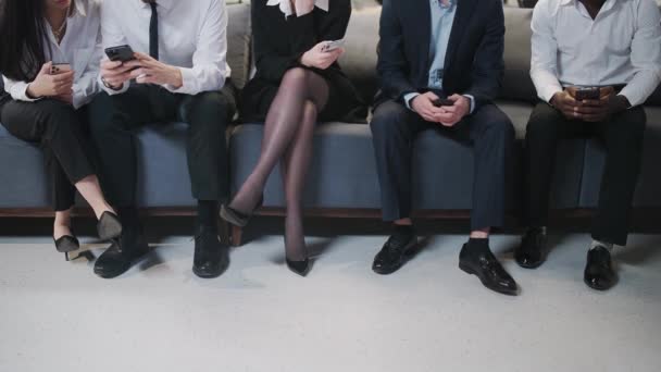 Група людей, які сидять в залі очікування і користуються смартфонами, офісна команда чекає на початок конференції в коридорі, вид на ноги, чоловіки і жінки сидять на стільцях . — стокове відео