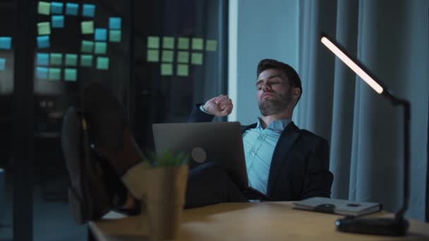 Trabajador de oficina joven trabaja hasta tarde, un hombre cansado con una computadora portátil en su regazo trabaja horas extras, trabajo nocturno, un hombre bosteza en el trabajo. — Vídeo de stock