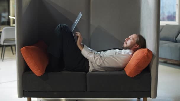 Administrador de oficina se encuentra en una silla grande y trabaja en un ordenador portátil, trabajo de oficina en un espacio abierto, un hombre escribe texto en un ordenador portátil, rutina. — Vídeos de Stock