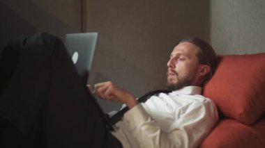 Ofis müdürü büyük bir sandalyede uzanıyor ve dizüstü bilgisayarda çalışıyor, ofis işi açık alanda, bir adam dizüstü bilgisayarına mesaj yazıyor, rutin bir iş..
