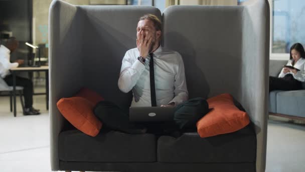 Office manager zit op een grote stoel en werkt op een laptop, kantoorwerk in een open ruimte, een vermoeide man typt tekst op een laptop, routine werk, man geeuwt op het werk. — Stockvideo