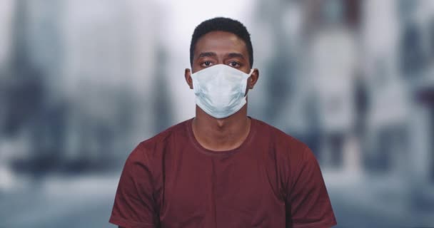 Afrikansk man med skyddande medicinsk mask tittar på kameran, porträtt av en man på en stad suddig bakgrund, skyddad från viruset. — Stockvideo