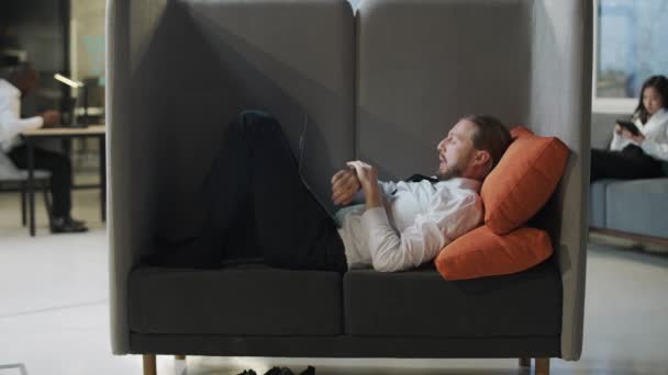 Zakenman ligt in een grote stoel en werkt op een laptop, kantoorwerk in een open ruimte, een man typt tekst op laptop, routine. — Stockvideo