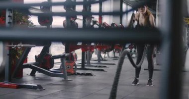 Spor salonundaki atletik kadın, savaş halatlarıyla egzersiz yapıyor, ağır çekimde çapraz dayanıklılık eğitimi alıyor..