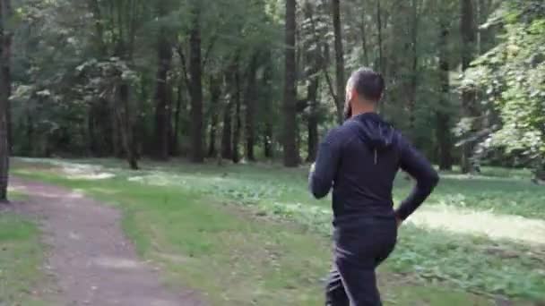 Hombre adulto corre en el parque, cámara siguiendo, corredor en auriculares inalámbricos, estilo de vida saludable en el bosque de verano. — Vídeos de Stock