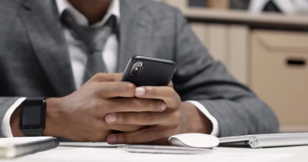 Modern bir ofiste çalışan, iş yerinde akıllı telefon kullanan genç bir Afrikalı adam, yakın plan, arka planda ofis içi.. — Stok video