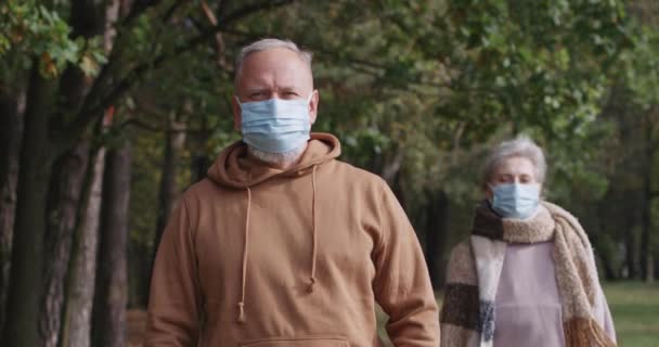Starší pár na procházce v lesoparku, podzimní den, šedovlasý muž a žena v lékařských maskách stojí v městském parku a dívají se do kamery, romantický okamžik. — Stock video