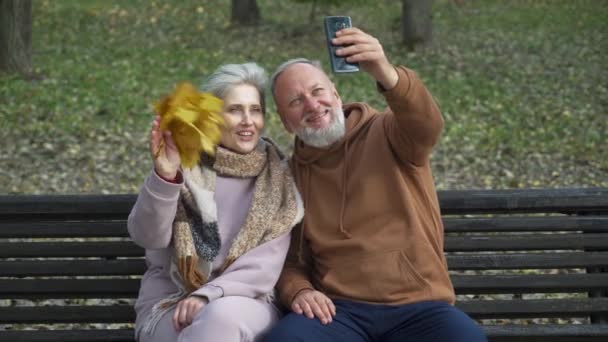 Ouderen echtpaar zit op een bank in een bos Park en neem een selfie video op een smartphone, grijs haar man en vrouw communiceren via conference call, oudere generatie. — Stockvideo