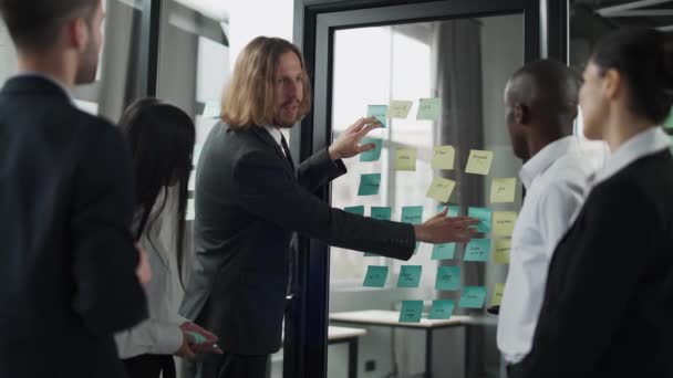 Business meeting, international management team at work, team is standing next to a glass board with paper stickers and notes, discussing work plans and planning financial work, brainstorm. — Stock Video