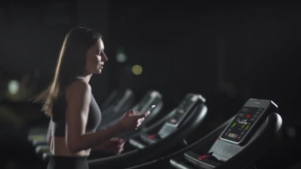 Kvinnlig idrottsman springer på löpband och använder en smartphone för att spåra sportaktivitet, aerob träning och uthållighetsträning i gymmet. — Stockvideo