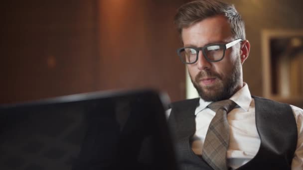 Jeune homme d'affaires en lunettes travaillant sur un ordinateur portable, homme en costume assis sur le canapé de l'hôtel, voyage d'affaires. — Video