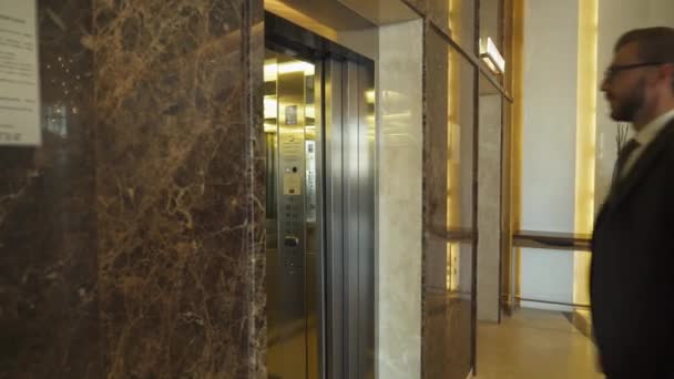 身穿西服的年轻人提着一个手提箱走下酒店大堂，男人走进电梯，开始了商务旅行. — 图库视频影像