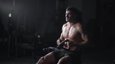 Spor salonundaki atletik adam kürek çekme makinesindeki blokları kaldırıyor, blok aleti ve spor aletleri üzerine eğitim alıyor, gece eğitimi alıyor, sinematik ışık alıyor..