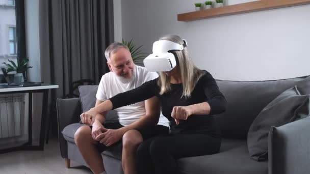 Älteres Paar entspannt sich auf dem Sofa im Wohnzimmer, eine erwachsene Frau mit Virtual-Reality-Brille spielt einen Motorrad-Fahrsimulator, Selbstisolierung — Stockvideo