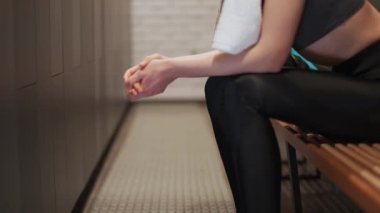 Genç bayan sporcu idmandan sonra dinlenir, kadın spor salonunun soyunma odasında bir bankta oturur ve terini havluyla siler..