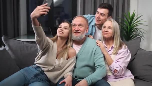 Alegre familia sentada en el sofá en la sala de estar, una chica se toma una selfie con sus padres y hermanos, niños y padres en un día familiar, la gente hace caras a la cámara. — Vídeo de stock