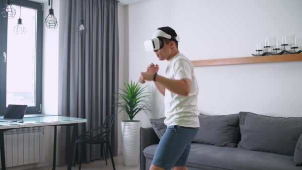 Młody człowiek w futurystycznych okularach wirtualnej rzeczywistości gra symulator gry walki, sport przy użyciu nowoczesnej technologii w samoizolacji, online szkolenia sportowe w salonie. — Wideo stockowe