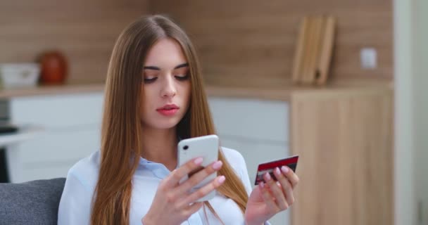 Jovem mulher está sentada no sofá na sala de estar e paga compras on-line usando um smartphone, interior da casa. — Vídeo de Stock