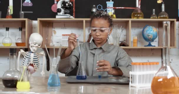 Laboratory experience in a chemistry lesson, african girl in protective glasses pours a blue liquid into a test tube, modern education at school, the girl looks at the tube of liquid. — Stock Video