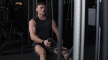 Spor salonundaki atletik adam kürek çekme makinesindeki blokları kaldırıyor, blok aleti eğitimi alıyor, sinematik ışıklandırma, 4k yavaş çekim..