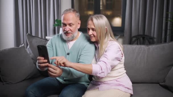 Pareja de ancianos se sienta en un sofá en la sala de estar y ver fotos en un teléfono inteligente, día de la familia, emociones positivas. — Vídeo de stock