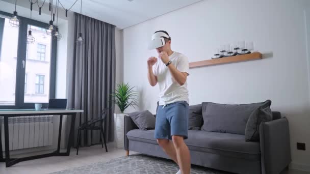 Młody człowiek w futurystycznych okularach wirtualnej rzeczywistości gra symulator gry walki, sport przy użyciu nowoczesnej technologii w samoizolacji, online szkolenia sportowe w salonie. — Wideo stockowe