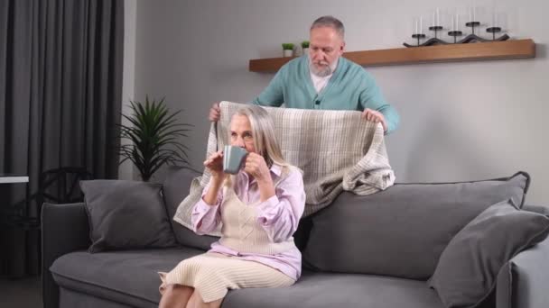 Äldre gråhåriga man täcker sin fru med en filt, tar hand om sina nära och kära, vuxna människor vilar bekvämt hemma, en äldre kvinna tittar på TV och dricker kakao sitter på — Stockvideo