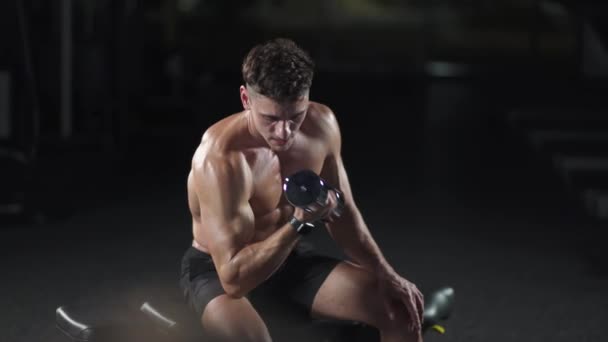 Hombre atlético entrena en el gimnasio, levanta pesas y realiza ejercicios para los brazos y bíceps, luz cinematográfica. — Vídeo de stock