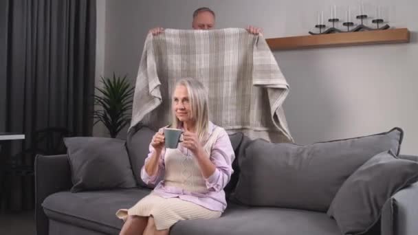 Äldre gråhåriga man täcker sin fru med en filt, tar hand om sina nära och kära, vuxna människor vilar bekvämt hemma, en äldre kvinna tittar på TV och dricker kakao sitter på — Stockvideo