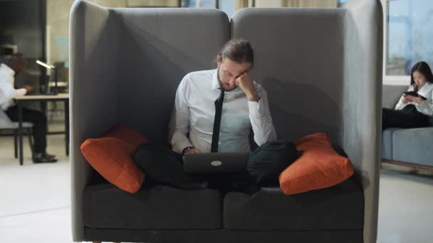 Kancelářský pracovník sedí na velké židli a pracuje v notebooku, kancelářská práce v otevřeném prostoru, unavený muž napíše text na notebook, rutinní práce, muž dokončí svou práci. — Stock video