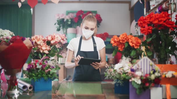 Fiorista donna in una maschera medica protettiva fa inventario in un negozio di fiori, una femmina utilizza una tavoletta schermo per contare il numero di fiori e piante in un negozio di fiori, lavorando in una pandemia. — Video Stock