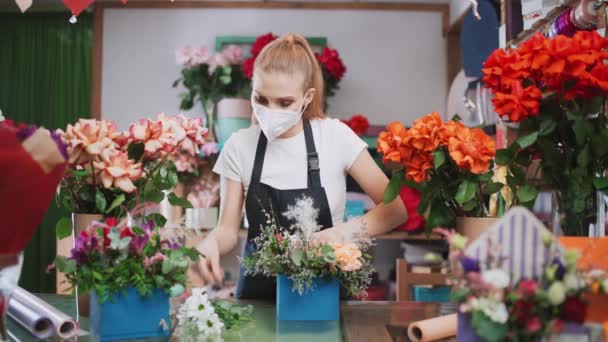 Giovane fiorista femminile raccoglie un mazzo di fiori in una scatola, donna allegra che lavora in un negozio di fiori, interno multicolore, lavora in una maschera protettiva nelle condizioni di una pandemia.. — Video Stock