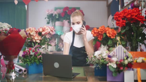 Florista mulher em uma máscara médica protetora discute a ordem, falando ao telefone e usando um laptop em uma loja de flores, trabalhando em uma pandemia. — Vídeo de Stock