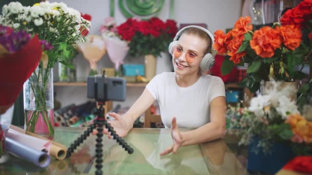 Fleuriste femelle dans les écouteurs communique avec le public par appel vidéo à l'aide d'un smartphone, fille streamer mène une diffusion en direct sur les réseaux sociaux dans les conditions de pandémie, auto-isolement dans — Video