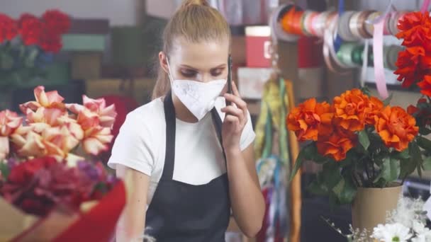 Florista mulher em uma máscara médica protetora discute a ordem, falando no smartphone e usando um laptop em uma loja de flores, trabalhando em uma pandemia. — Vídeo de Stock