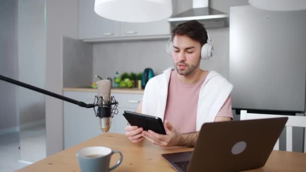 Jovem streamer faz um podcast ao vivo para redes sociais, trabalhando remotamente enquanto está sentado na cozinha, usando laptop e tablet de tela, trabalhando no contexto da pandemia do coronavírus — Vídeo de Stock