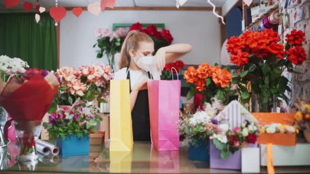 Floristería femenina en máscara médica protectora empaca cajas con plantas y regalos en bolsas de papel de colores, mujer alegre trabajando en una tienda de flores y plantas, trabajando en un entorno pandémico. — Vídeo de stock