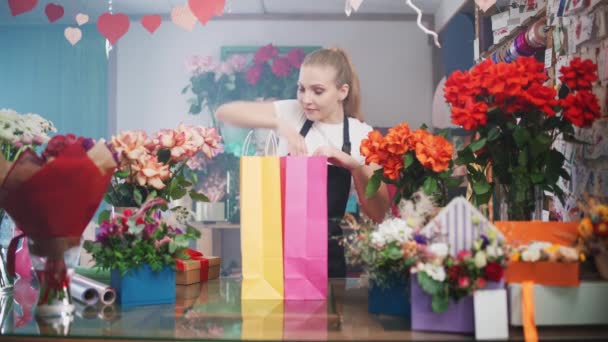 Neşeli kadın çiçekçi, içinde bitkiler ve renkli kağıt torbalarda hediyeler olan kutular paketliyor. Çiçek ve bitki dükkanında çalışan genç bir kadın.. — Stok video