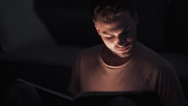 Portret młodego mężczyzny siedzącego w domu na podłodze i czytającego książkę, ciepłe światło, samoizolacja podczas pandemii, mistyczny efekt świetlny. — Wideo stockowe