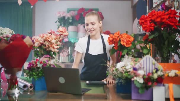 Female florist discusses the order by talking on video call, using a laptop and screen tablet in a flower shop, working online, the female chooses flowers for the customer. — Stock Video