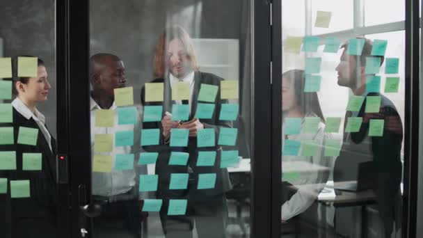 Obchodní jednání, mezinárodní management tým v práci, tým stojí vedle skleněné tabule s papírovými samolepkami a poznámkami, diskutovat o pracovních plánech a plánování finanční statistiky, brainstorm. — Stock video